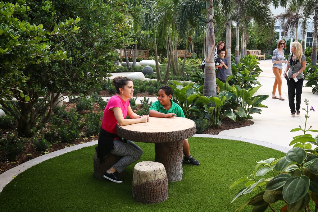 In a nook nestled within lush planting and encircled by a distinctive paving band, a teacher and child sit at a table and stools constructed of rustic logs, smiling and chatting.