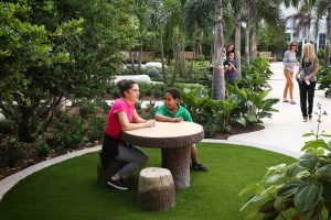 In a nook nestled within lush planting and encircled by a distinctive paving band, a teacher and child sit at a table and stools constructed of rustic logs, smiling and chatting.