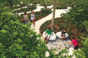 Seen from above, two children sit on large smooth rocks nestled in the greenery, talking to a seated adult. In the background, three more adults stand and are talking.