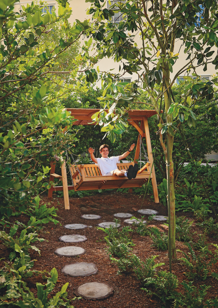 A young person sits on a swinging bench, smiling with their hands waving with joy in the air.