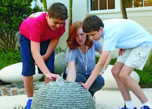 A teacher and two young people crouch down to touch a stone water sphere, with a small fountain of water on the top and ribbed sides, providing both proprioceptive and vestibular sensory experiences. In the background, there are also smooth rocks that offer varied tactile engagement.
