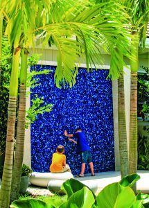 Viewed through the palm trees, a child stands with both of their hands pressed into the water wall, a large blue feature wall offering sensory opportunities, while another looks on from their seat on a nearby rock.