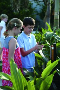 Two children stand at a musical sculpture while one holds the rubber mallet for the instrument. They are surrounded by foliage. In the background, another child can be seen exploring the area.