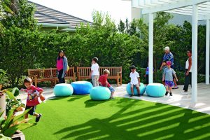 In an open green space, several children play on large blue cushions while adults mill around. Behind this area are two large, curved benches in front of tall green hedging. A pergola provides some shade.